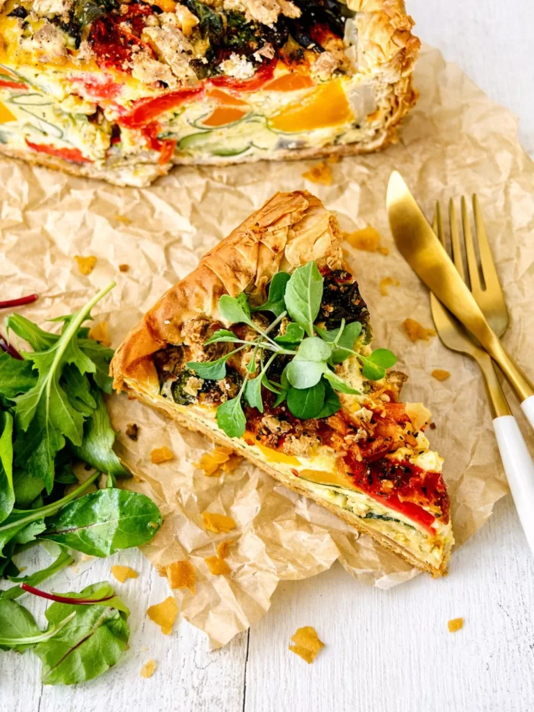 Slice of deep-dish roasted vegetable quiche on crinkled baking paper, showing layers of pumpkin, zucchini and red capsicum in creamy filling, topped with microgreens, with a small side of leafy salad and gold cutlery on a white timber surface.