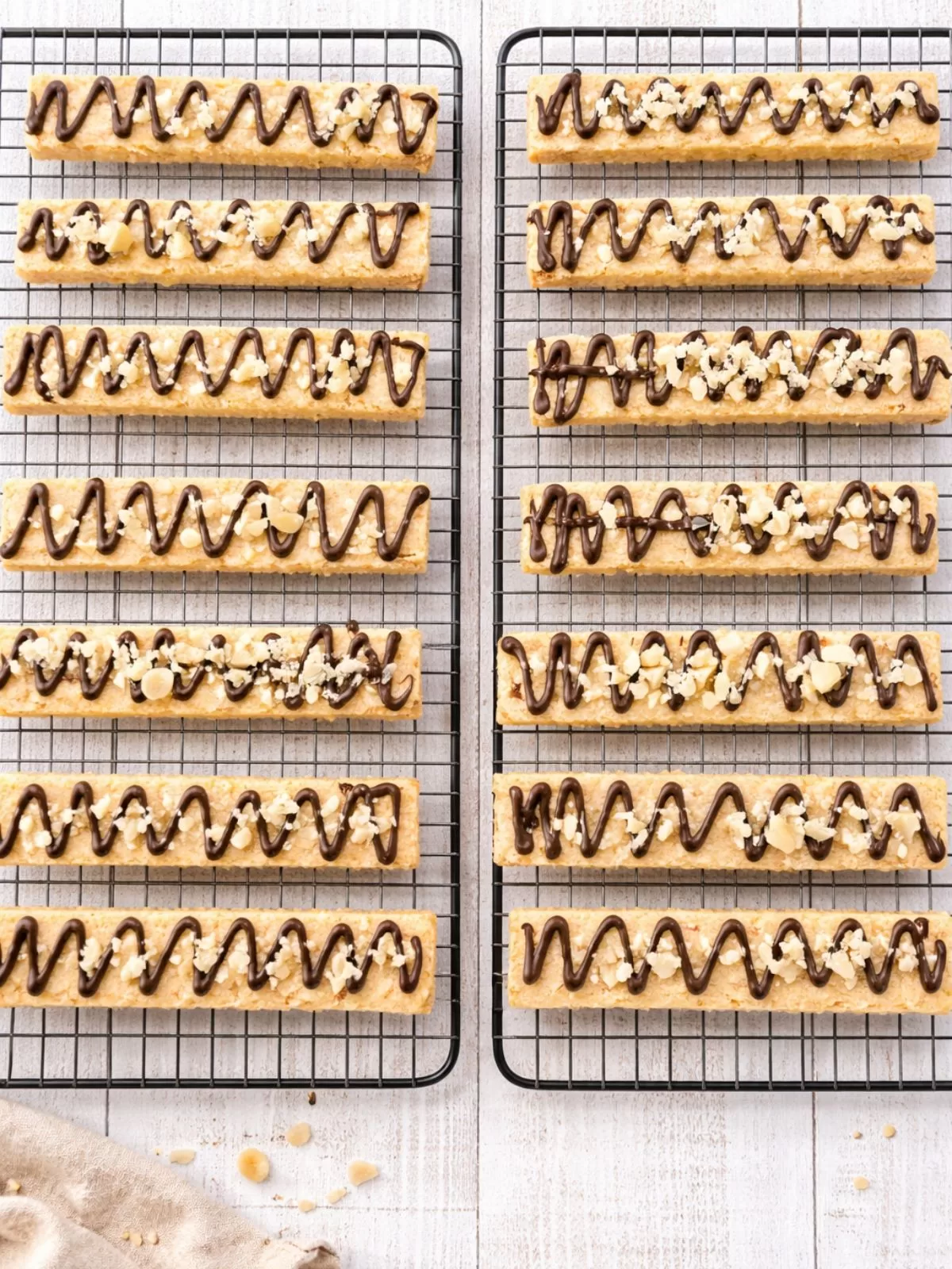 Overhead flat lay of macadamia nut shortbread fingers arranged on cooling racks, drizzled with dark chocolate and sprinkled with chopped macadamias, set on a white timber background.