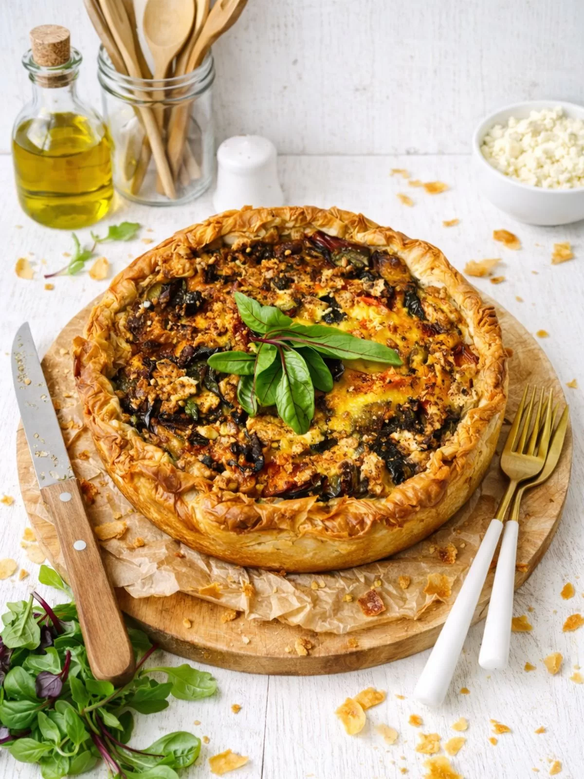Close-up of a slice of deep-dish roasted vegetable quiche with flaky golden filo pastry, showing layers of roasted pumpkin, zucchini and creamy egg filling, topped with fresh greens, sitting on baking paper with the rest of the quiche softly blurred in the background.