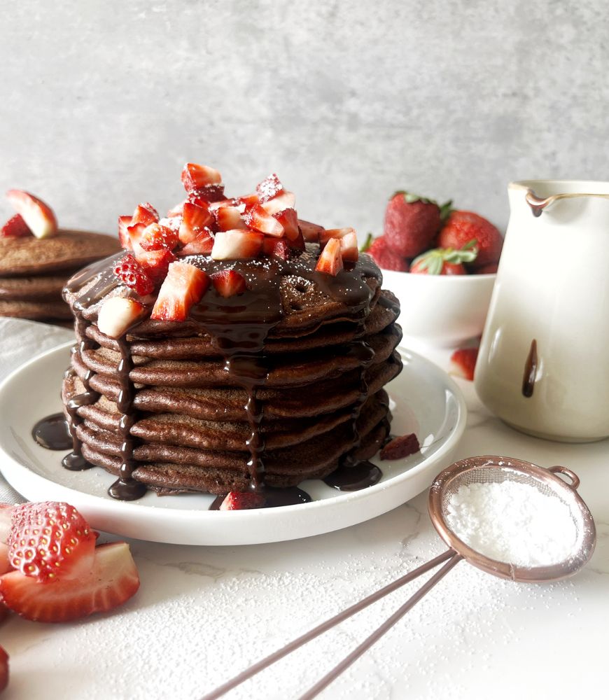 Chocolate pancake stack on white plate drizzled with chocolate sauce and chopped strawberries