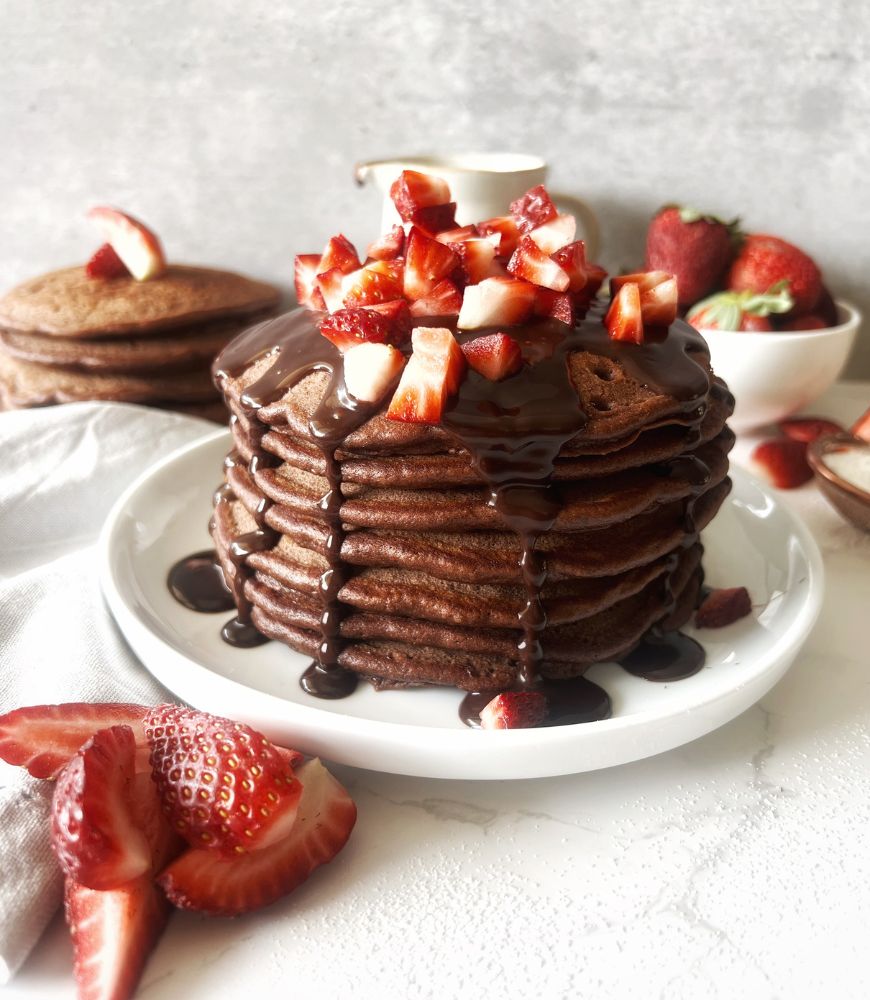 chocolate pancakes stacked on white plate with chopped straweberries and chocolate sauce
