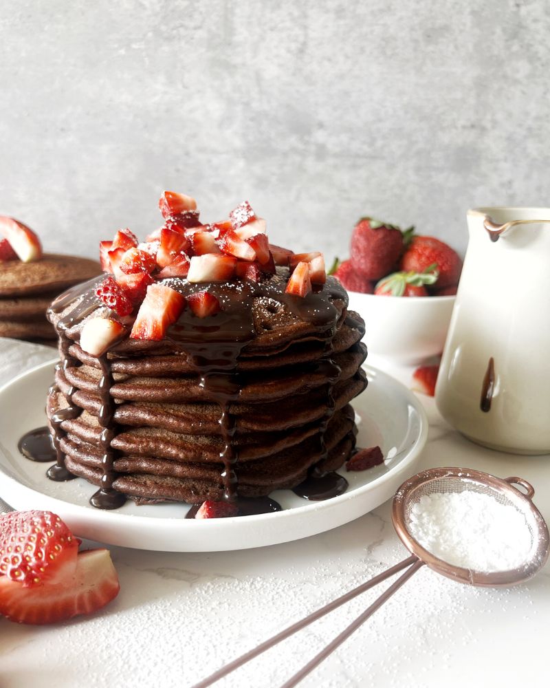 chocolate pancakes stacked on a white plate with chocolate sauce and strawberries on top