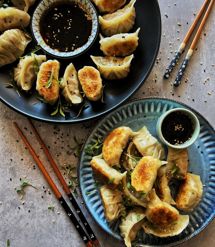 CRISPY PORK DUMPLINGS ON 2 PLATES WITH BLACK SESAME DIPPING SAUCE. CHOPSTICKS ON EITHER SIDE AND SESAME SEEDS AND MICRO-GREENS TO GARNISH