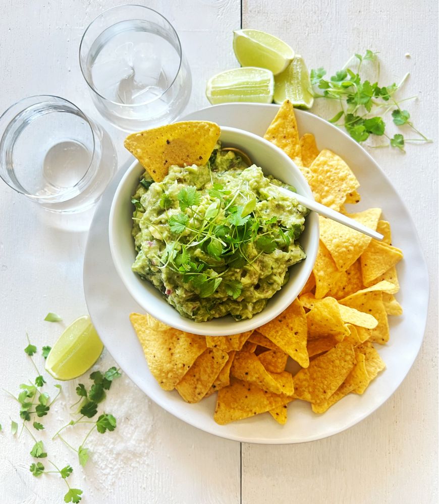 Easy Guacamole in a white bowl on a white plate with corn chips on a white wood background. 2 glasses of water, lime wedges and coriander scattered around the plate