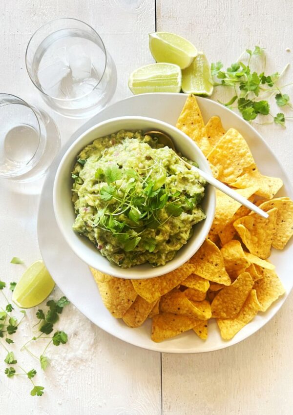 Guacamole in white bowl sitting on a white plate with corn chips around it. 2 glasses of water and some herbs sprinkled around it