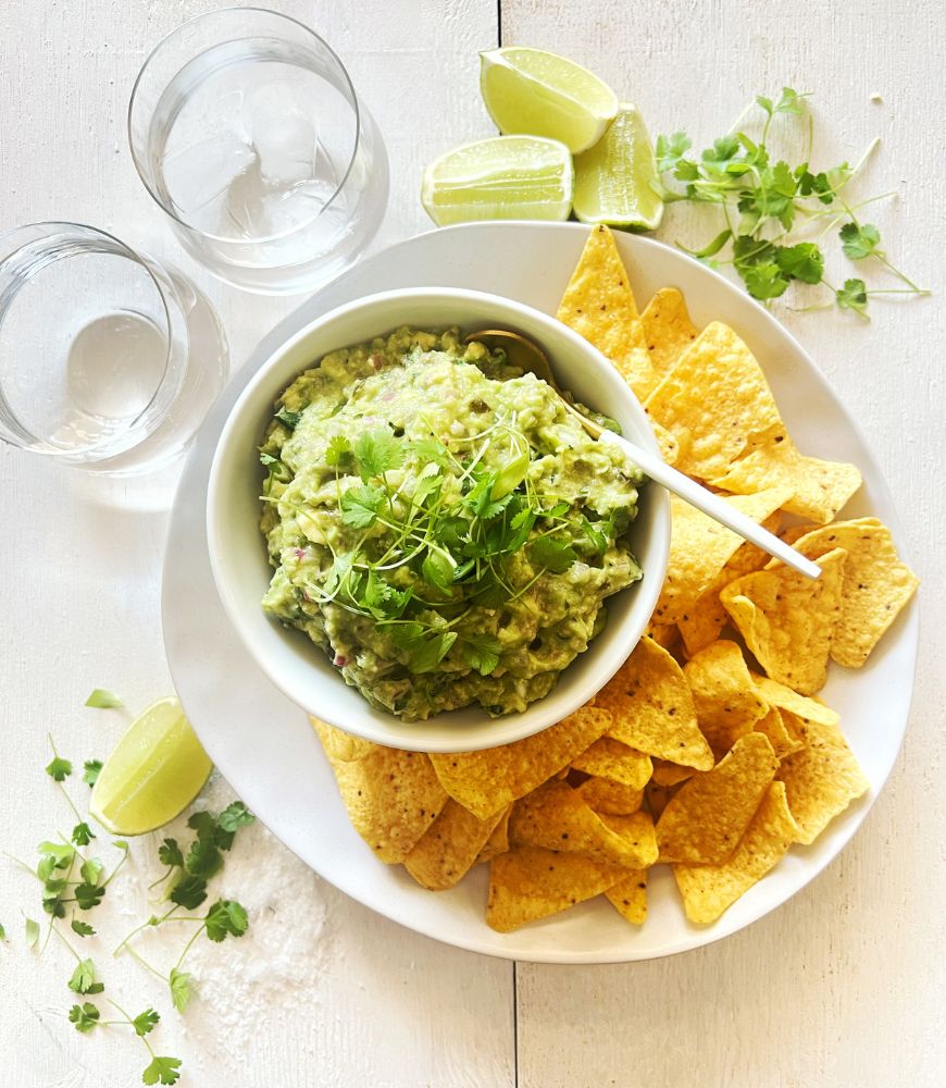 Easy 5 Ingredient Guacamole in a white bowl on a white plate with corn chips on a white wood background. 2 glasses of water, lime wedges and coriander scattered around the plate