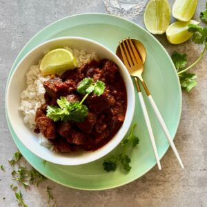 Mexican Chilli Beef in white bowl on a green plate. Lime wedges and coriander to the sides