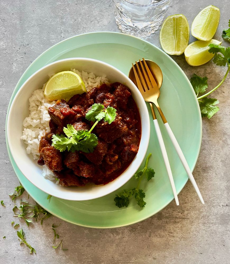 Mexican Chilli Beef in white bowl sitting on a green plate on a grey background. lime wedges and coriander around the plate