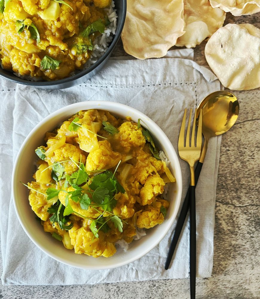 cauliflower curry in a bowl with spoon and fork. poppadums in the background