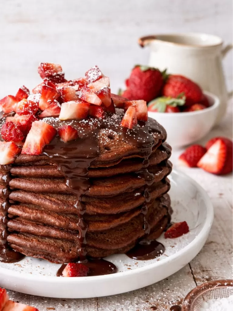 Close-up of chocolate buttermilk pancakes stacked on a white plate, dripping with glossy chocolate ganache and topped with chopped strawberries and a light dusting of icing sugar, set on a white timber background with a small jug of ganache and fresh strawberries softly blurred behind.