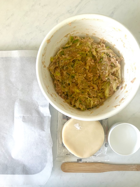 Pork dumpling mixture in a white bowl with baking tray, dumpling wrappers, water and spatula next to it