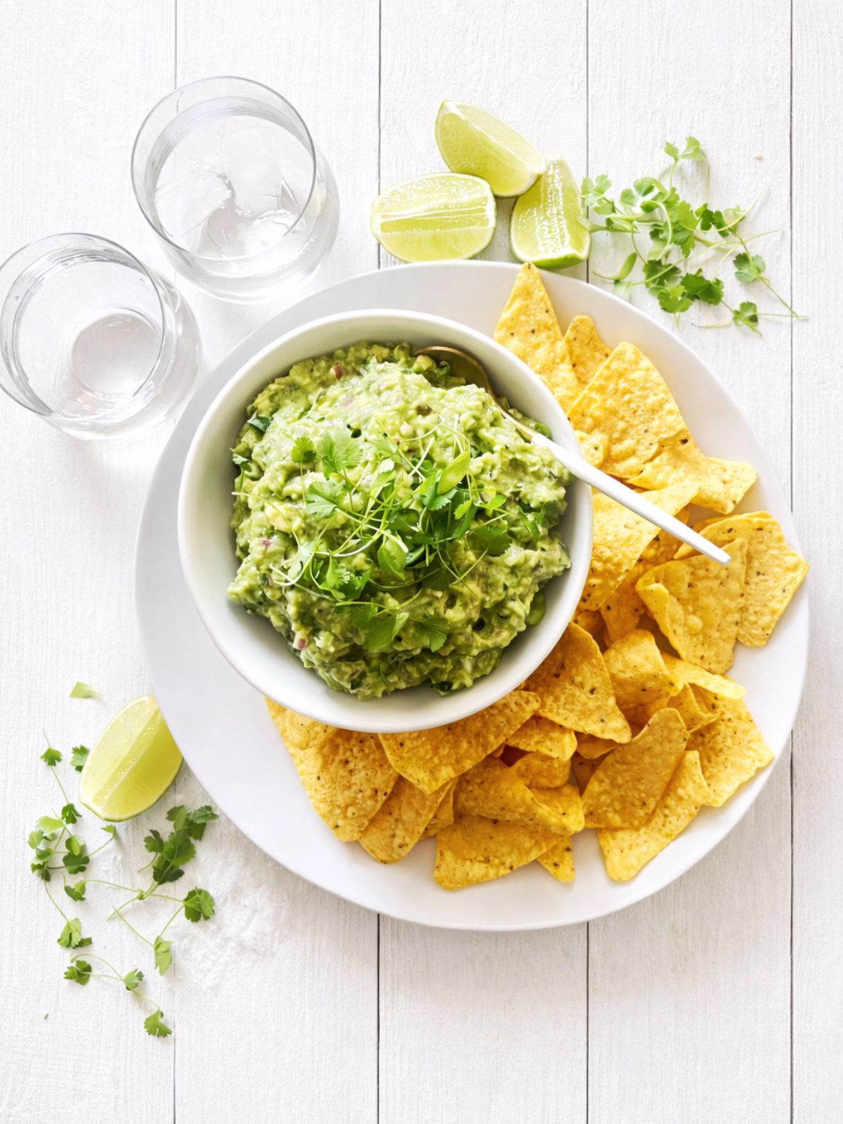 Bowl of chunky guacamole topped with fresh herbs, served with corn chips on a white plate over a white timber background with lime wedges and glasses of water.