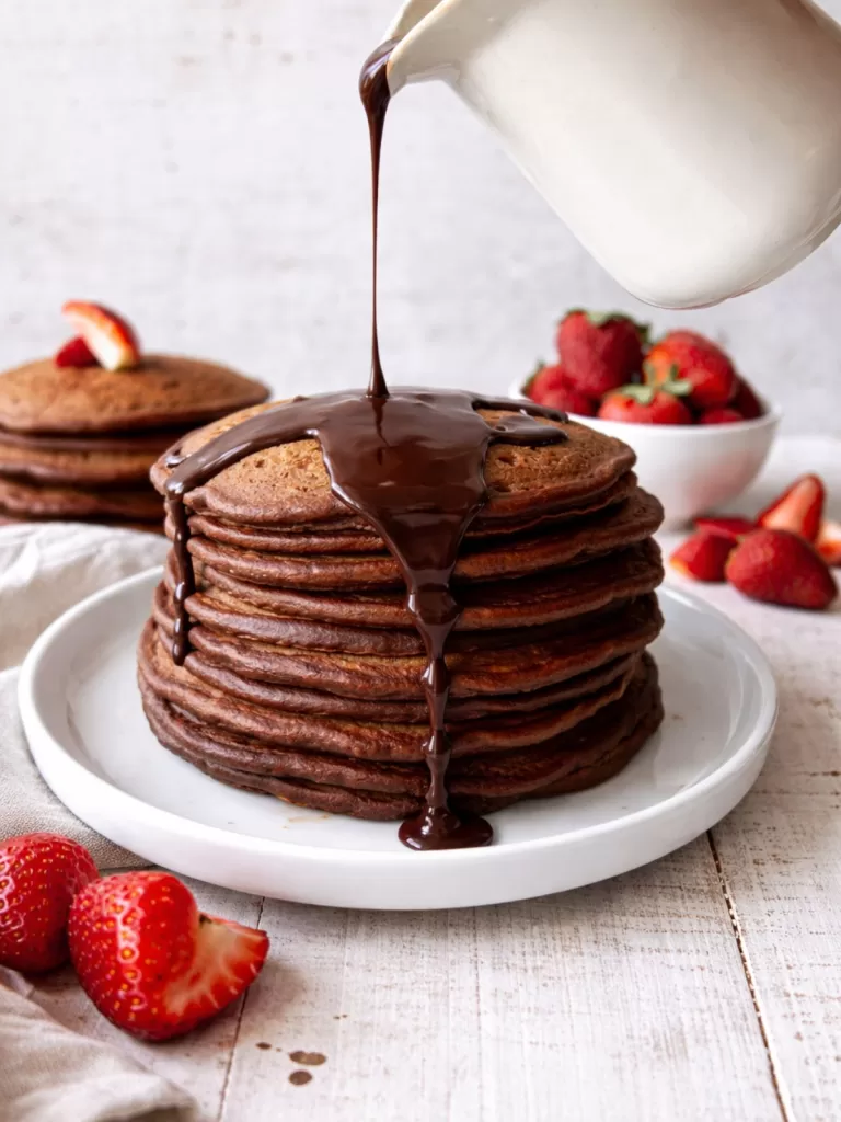 Stack of chocolate buttermilk pancakes on a white plate with glossy chocolate ganache being poured over the top, set on a white timber background with fresh strawberries and a small bowl of berries in soft focus behind.