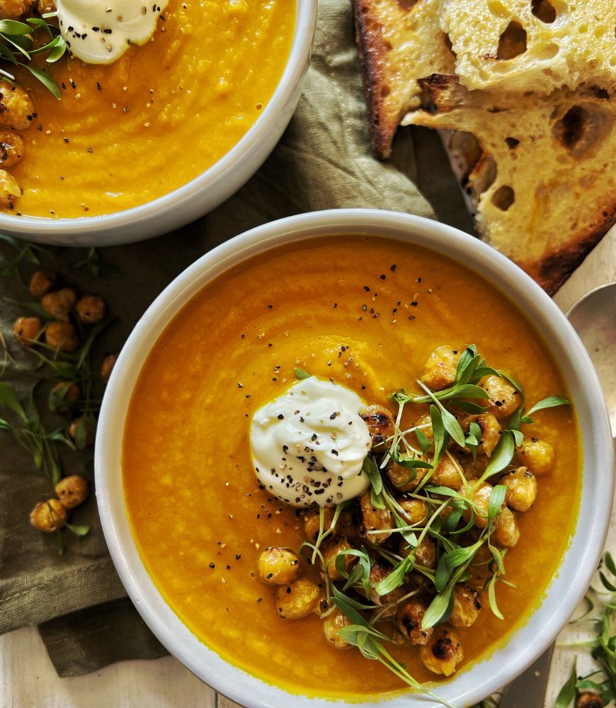 2 white bowls filled with carrot soup . Sourdough toast to right side of bowls and silver soup spoon. Soup garnished with toasted chickpeas, yoghurt and micro-greens.