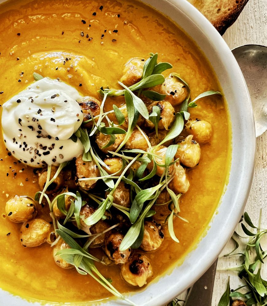 close up of carrot soup in bowl topped with yoghurt, spiced chickpeas and micro-greens.