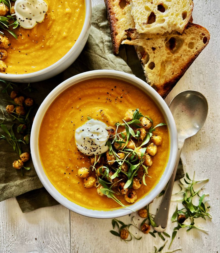 2 white bowls filled with roasted carrot soup with chickpeas. Sourdough toast to right side of bowls and silver soup spoon. Soup garnished with toasted chickpeas, yoghurt and micro-greens.