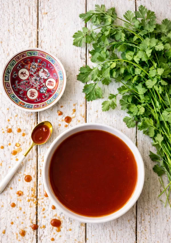 Overhead flat lay of a bowl of glossy sweet and sour sauce on rustic white timber slats, with scattered sauce droplets, a gold spoon with sauce, a small decorative Chinese dish, and a fresh bunch of coriander in natural light.