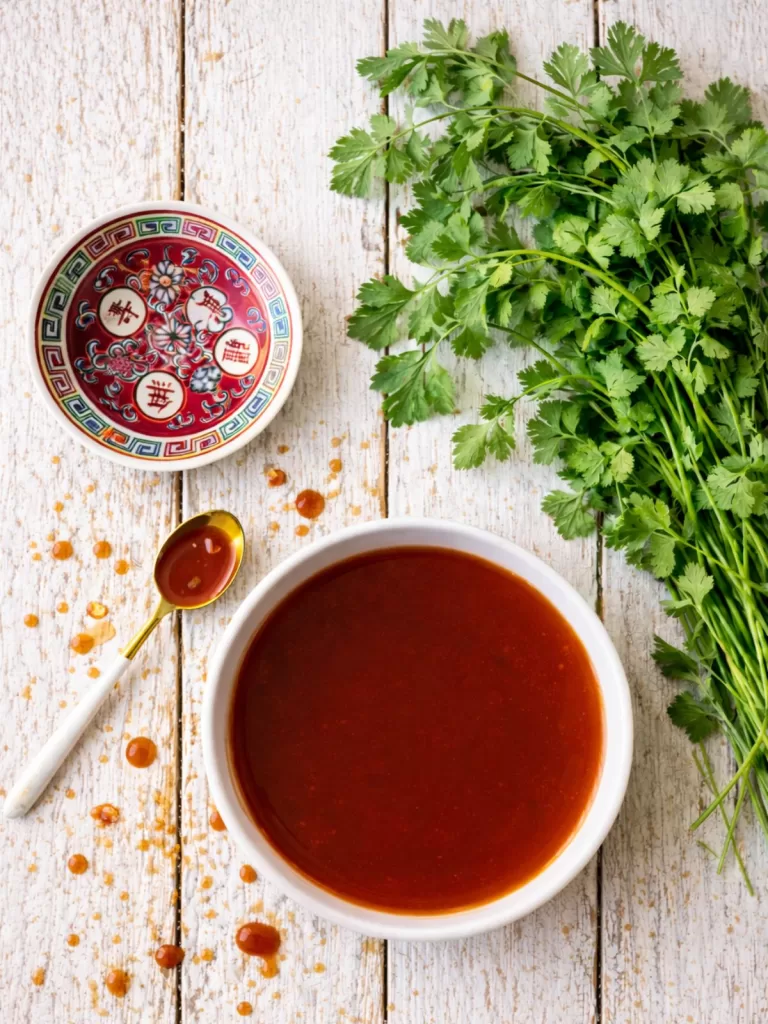 Overhead flat lay of a bowl of glossy sweet and sour sauce on rustic white timber slats, with scattered sauce droplets, a gold spoon with sauce, a small decorative Chinese dish, and a fresh bunch of coriander in natural light.