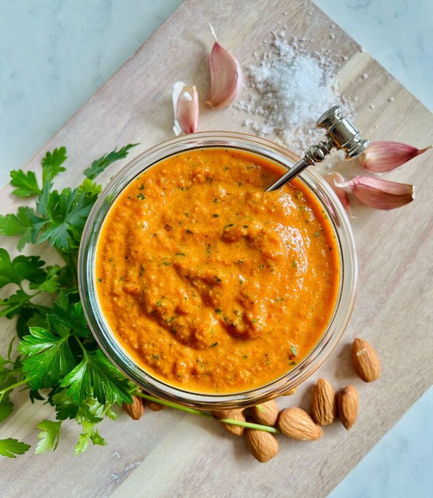 Easy Romesco Sauce in a glass jar with parsley, garlic, salt and almonds around it.