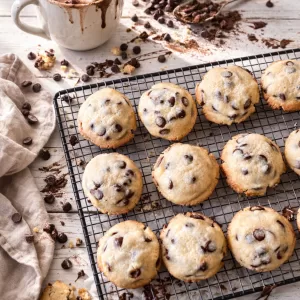Chocolate chip cookies cooling on a wire rack over a white timber table, with a mug of hot chocolate, a tipped measuring cup spilling chocolate chips, crumbs, and a linen napkin in natural light.