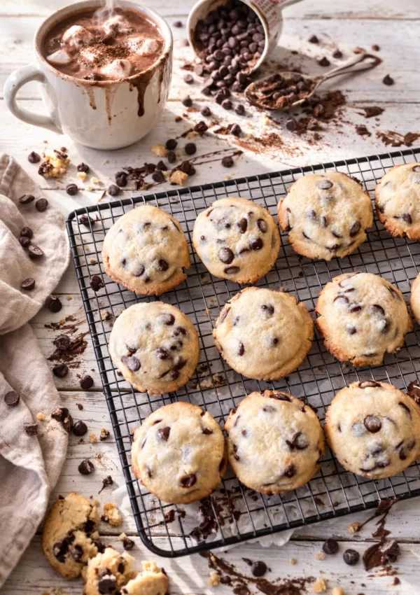 Chocolate chip cookies cooling on a wire rack over a white timber table, with a mug of hot chocolate, a tipped measuring cup spilling chocolate chips, crumbs, and a linen napkin in natural light.