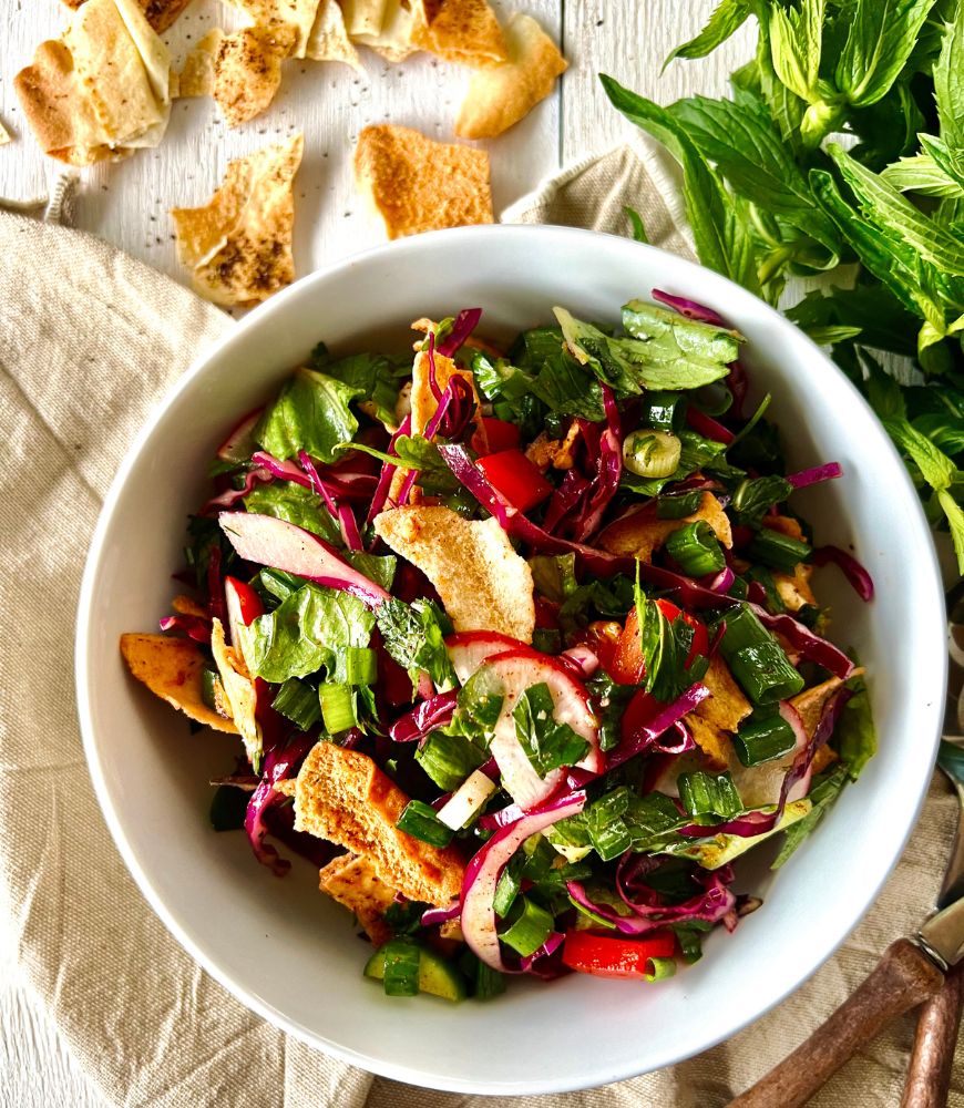 fattoush salad in white bowl on white wooden background. Beige napkin under the bowl. Salad servers, mint and pita crisps around the bowl.