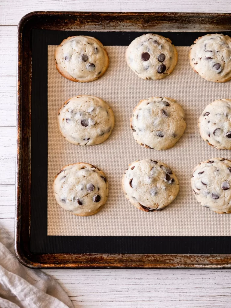 Freshly baked chocolate chip cookies on a lined baking tray over a white timber background, photographed from above in natural light.