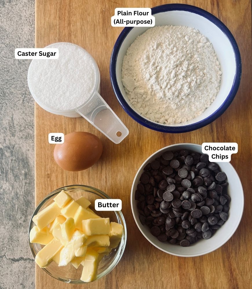 Ingredients on a wooden board in small bowls. Sugar, flour, egg, butter and choc chips