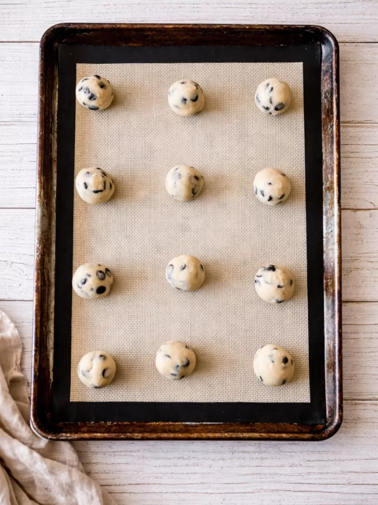 Chocolate chip cookie dough balls evenly spaced on a lined baking tray over a white timber background, shot from above in natural light.