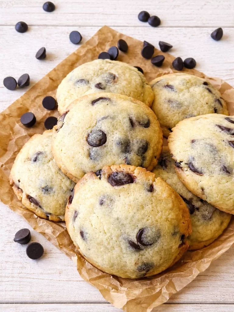 Stack of chocolate chip cookies on scrunched rustic baking paper over a white timber surface, with scattered chocolate chips around.
