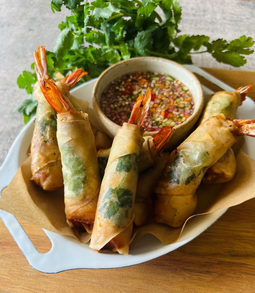 Thai prawn and pork spring rolls in white dish sitting on wood board with dipping sauce and coriander in background