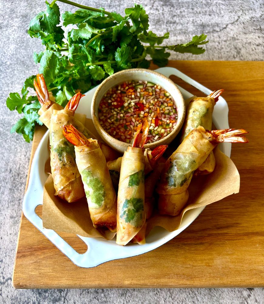 Thai spring rolls in white dish sitting on wood board with dipping sauce and coriander in background