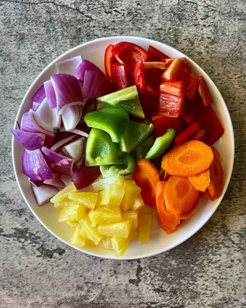 white plate with chopped red onion, red capsicum, green capsicum, carrot and pineapple pieces on it. Marble background