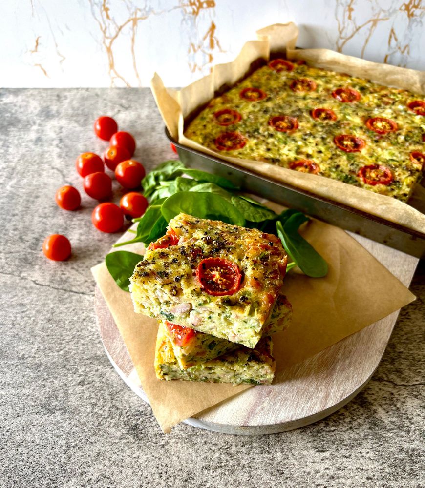 zucchini and bacon slice stacked on baking paper on board with slice in background. Tomatoes and spinach leaves scattered around