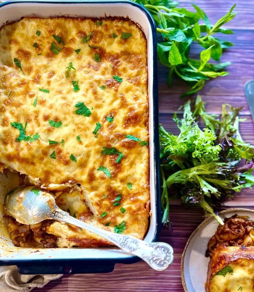 pasta bake in white and black dish with salad and herbs around it. Silver spoon in it on a wooden style background