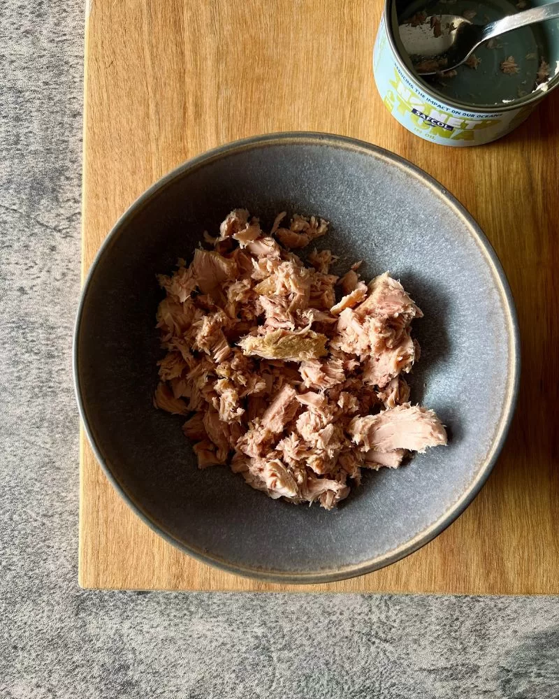 tuna in a grey bowl sitting on top of wooden board with empty can of tuna behind it