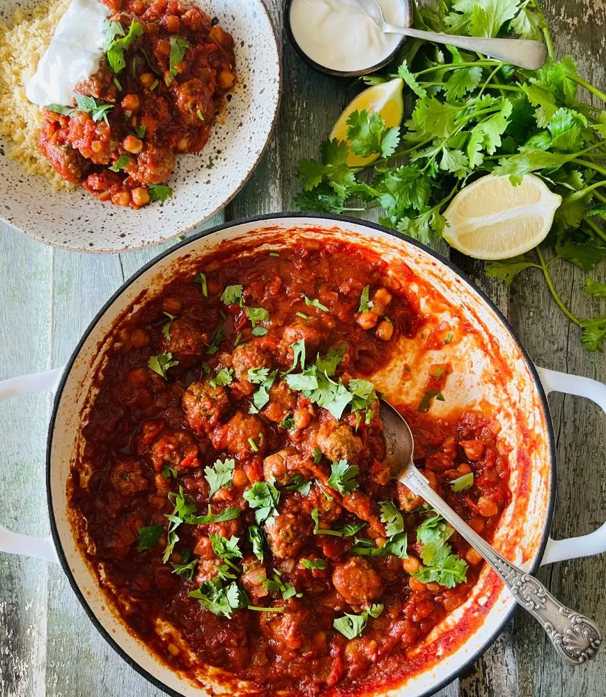 white cast iron dish filled with a tomato based meatballs, plate behind it with the meal and coriander, lemon wedges and yoghurt around it