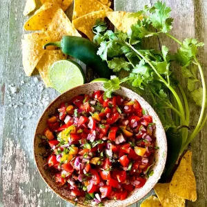 Fresh tomato salsa in speckled dish on green wood background. Corn chips, coriander, limes and jalapenos around it.