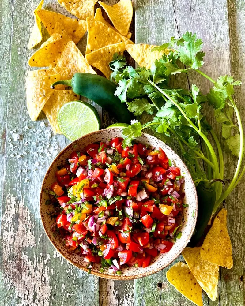 Fresh tomato salsa in speckled dish on green wood background. Corn chips, coriander, limes and jalapenos around it.