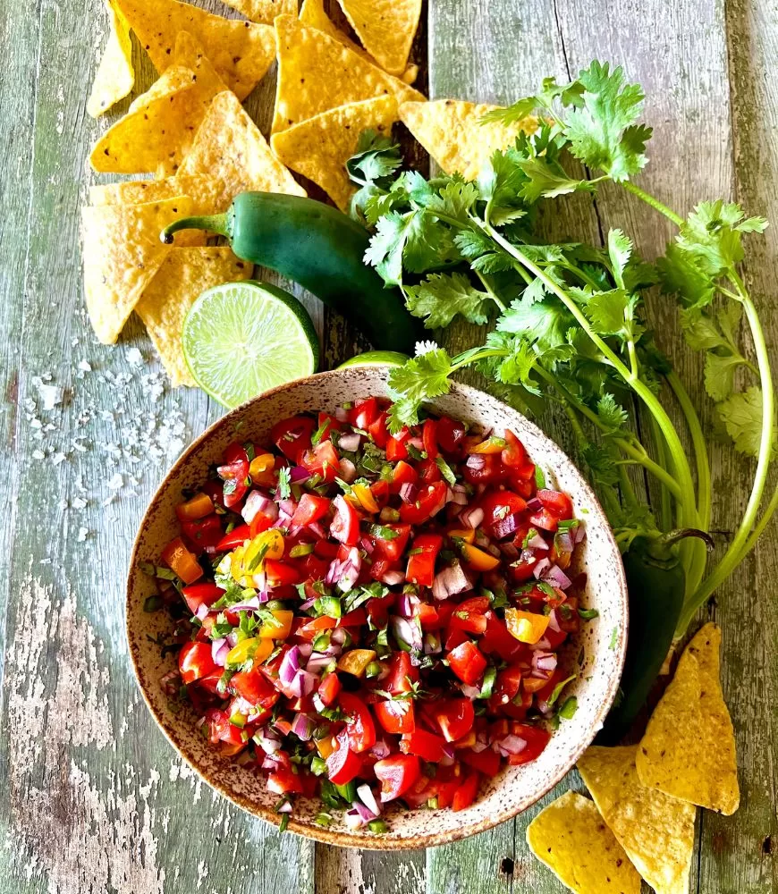 Fresh tomato salsa in speckled ish with fresh coriander, lime halves, corn chips around it on green washed out wood background