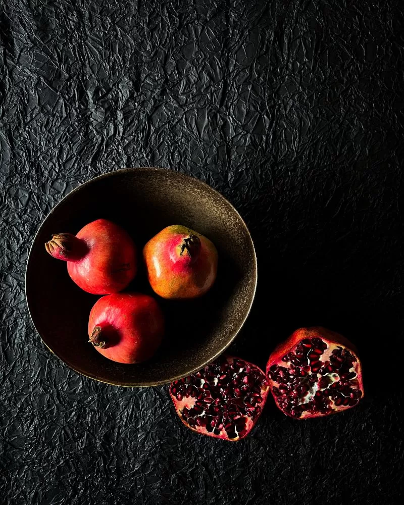 pomegranates in a brown bowl on a black background. Open cut one next to the bowl.