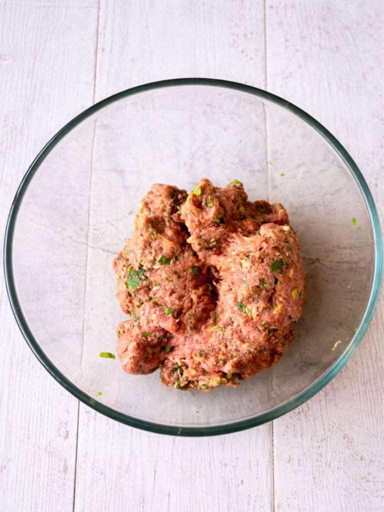 Glass bowl with seasoned lamb mince mixture for meatballs on a white timber background.