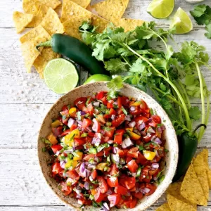 Overhead photo of fresh pico de gallo in a speckled ceramic bowl on a rustic white timber table, surrounded by tortilla chips, a whole jalapeño, fresh coriander, olive oil, pink salt and lime wedges in natural light.