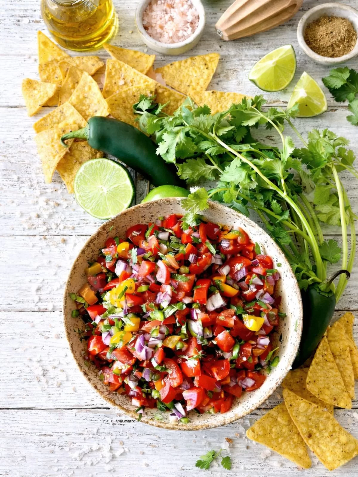Overhead photo of fresh pico de gallo in a speckled ceramic bowl on a rustic white timber table, surrounded by tortilla chips, a whole jalapeño, fresh coriander, olive oil, pink salt and lime wedges in natural light.