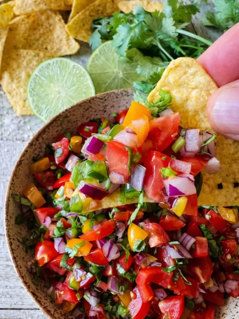 Close-up of a tortilla chip scooping fresh pico de gallo from a speckled ceramic bowl on a rustic white timber table, with chopped tomatoes, red onion, jalapeño and coriander visible, and tortilla chips and lime slices blurred in the background.