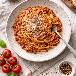 Overhead view of spaghetti bolognese in a bowl on a white wooden table with parmesan and a fork.
