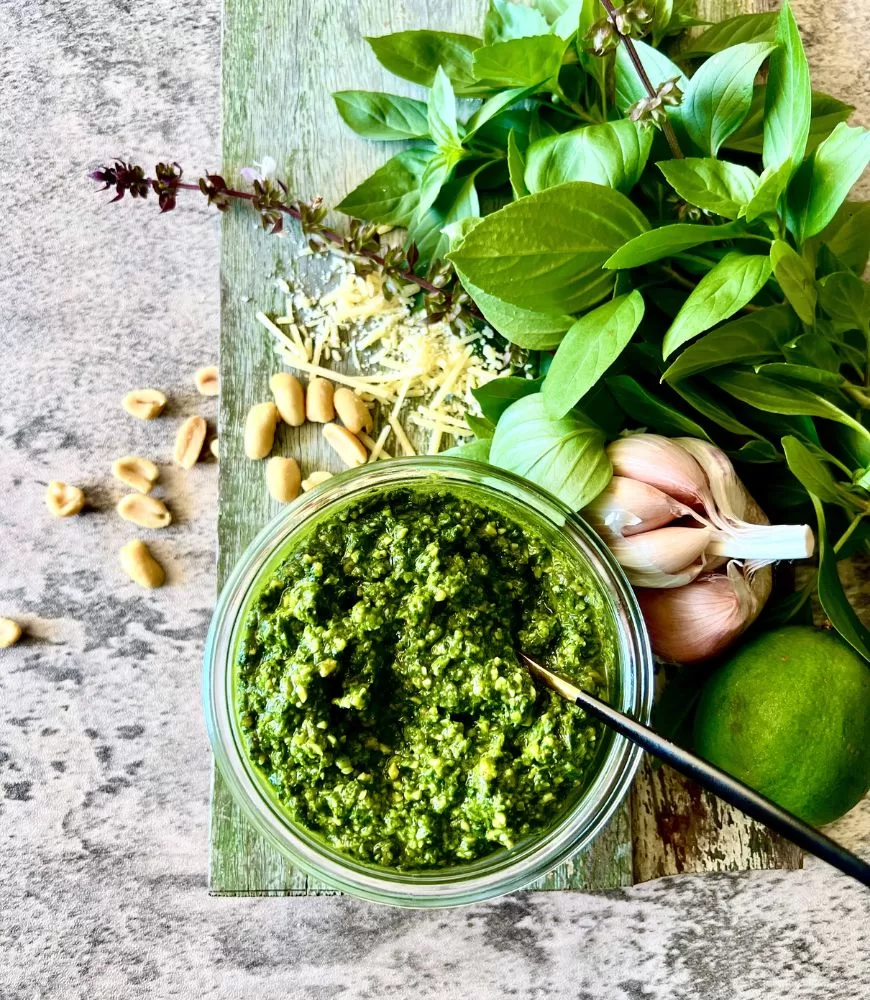 Thai basil pesto in glass jar on a board with peanuts, parmesan, garlic, Thai basil and lime. Black spoon in the jar