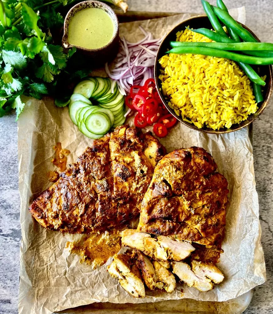 tandoori chicken breast on baking paper on a tray with bowl of yellow rice, beans, cucumber, chilli, onion, sauce and corinader