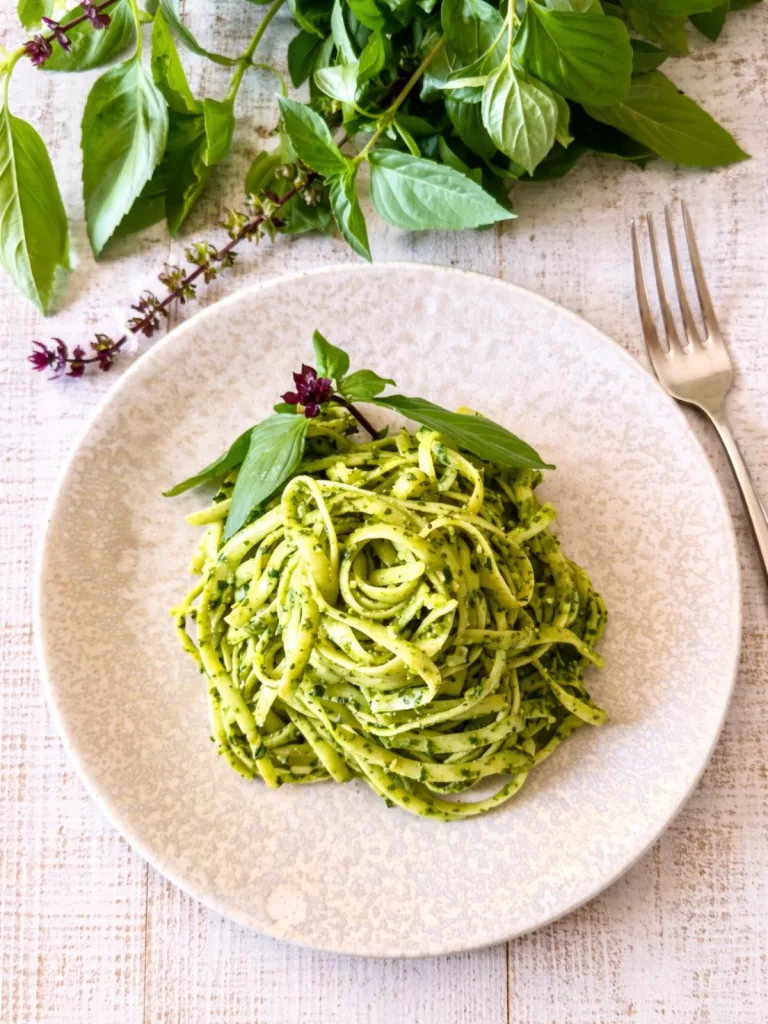 Plate of linguine tossed in Thai basil pesto on a white timber background, garnished with fresh basil leaves and served with a fork in bright natural light.