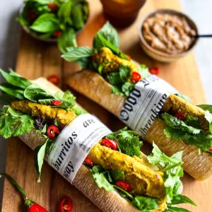 2 Thai chicken satay baguettes on a board with herbs, water, and satay sauce